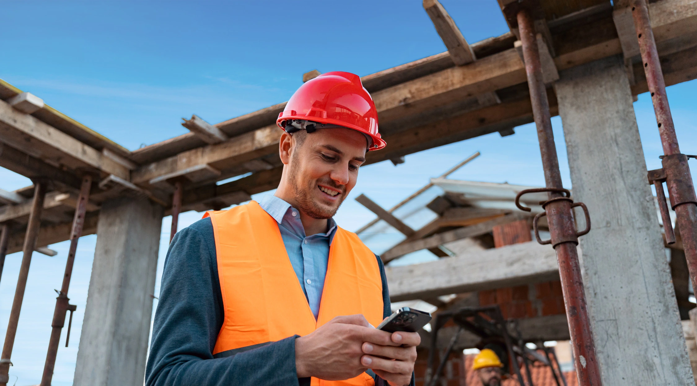 Man in high visibility vest and hardhat looking at phone while on a construction site.